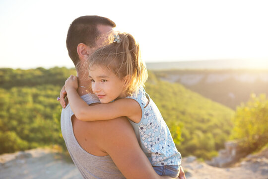 The Father And Daughter Hug Tightly, The Girl Holds The Father By The Neck, The Father - In His Arms. Father's Day, Strong Feelings, Family. Summer, Sunshine, Greenery, Mountains, Travel