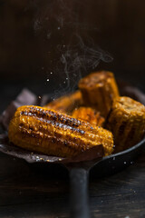 Grilled corn on the cob on black cooking pan with salt in the air. Wooden rustic table,  dark background. Grilled vegetables, vegetarian food, bbq. Copy space, dark mood photography.