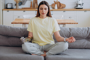 Woman with a bionic prosthetic arm in a yellow T-shirt sits at home on the sofa in the lotus position and meditates with her eyes closed, enjoying the peace and quiet - obrazy, fototapety, plakaty