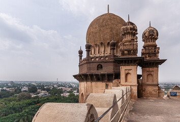 Vijayapura, Karnataka, India - November 8, 2013: Gol Gambaz Mausoleum. Horizontal view on brown stone top of one tower under light blue cloudscape. Cityscape and green foliage down there. © Klodien
