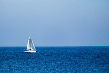 A white sailboat in the middle of the calm sea and under a wide clear blue sky.