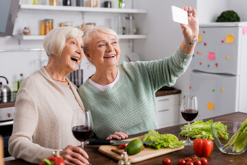 cheerful senior woman taking selfie with retired friend in kitchen