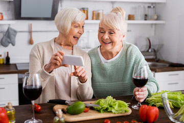cheerful senior woman pointing with finger at smartphone near friend and vegetables on table