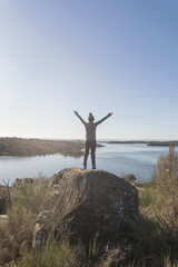 Fototapeta premium Young and attractive female with her arms opened and located on top of a rock with a lake in front of her during a sunny and cold winter morning.