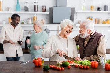 happy senior man and woman cooking salad near multiethnic friends on blurred background