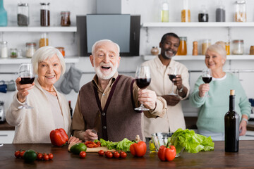 amazed senior man and woman holding glasses of wine near multicultural friends on blurred background