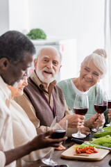 happy retired man smiling while holding glass of wine near multicultural friends on blurred foreground
