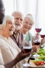 cheerful interracial pensioners laughing while holding glasses with wine