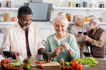 African american man and happy senior woman preparing salad near retired friends on blurred background - Powered by Adobe