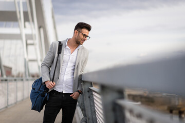 Happy young businessman standing on the bridge. Yuppie outdoor holding bag. Smart young professional walking outside