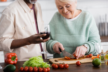 happy senior woman cutting ripe bell pepper near african american husband with glass of wine