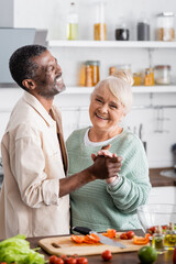 senior multicultural husband and wife smiling while holding hands in kitchen