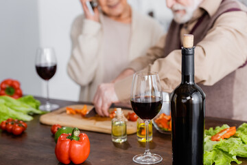 bottle with red wine near glass, vegetables and retired couple on blurred background