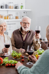 senior interracial friends holding glasses with wine and smiling near vegetables on table