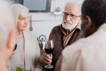 senior man in eyeglasses holding glass of red wine near retired multiethnic friends