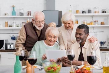 senior multicultural people looking at digital tablet with happy friends near tasty food on table