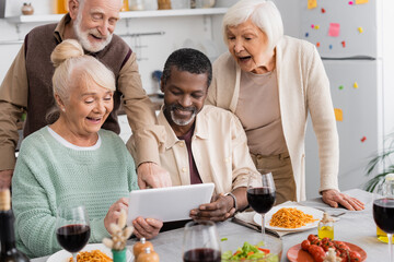 retired multicultural people looking at digital tablet with happy senior friends near tasty food on table