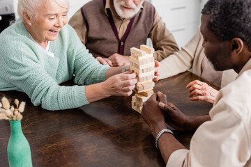 happy multicultural pensioners playing tower wood blocks game at home