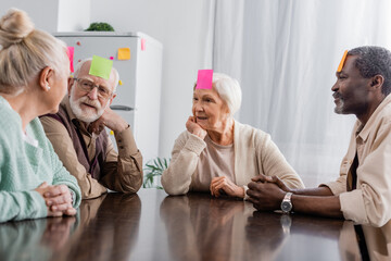 Obraz na płótnie Canvas multicultural senior friends with sticky notes on foreheads playing game in kitchen