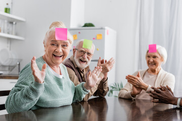 excited and multicultural senior friends with sticky notes on foreheads playing guess who game in kitchen