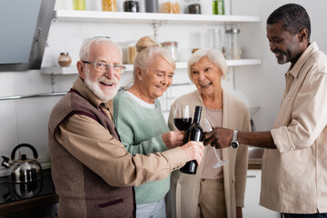 happy retired man holding bottle with wine near senior multicultural friends in kitchen