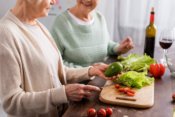 cropped view of senior woman holding knife and avocado near retired friend in kitchen
