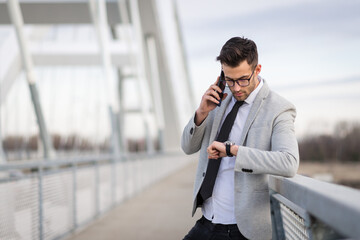Young businessperson standing on the bridge talking on smartphone and checking the time. Yuppie or young entrepreneur outdoors relaxing