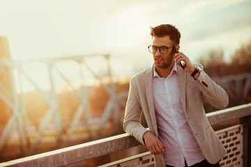Young businessman crossing the bridge at sunset talking on smartphone. Yuppie outdoors talking on mobile and smiling