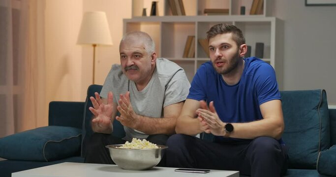 Father and son are watching a sports match of their favorite team on TV, sitting on the couch. They clap their hands approvingly and raise their hands enthusiastically after a goal has been scored.