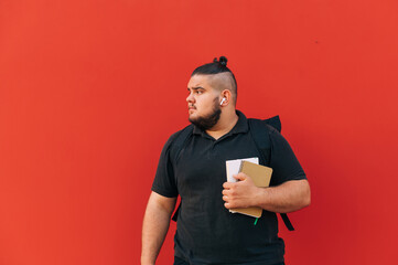 Serious young man in casual clothes and wireless headphones and with books in his hands stands on a background of a red wall and looks away.