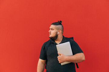 Handsome charismatic man with laptop in hands and wireless headphones looking to the side on red...