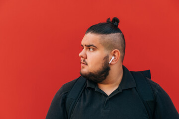 Bearded overweight man in wireless headphones looking to the side, close portrait on red background.