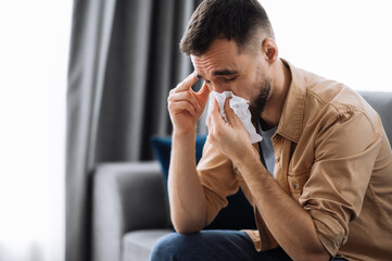 Unhealthy young man. Caucasian guy is sick with flu, he sneezes and blows his nose in a napkin while sitting on the couch at home