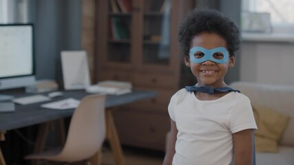 Panning slowmo waist-up portrait of happy cute mixed-race boy in blue superhero mask and cloak standing in bright modern living room looking at camera smiling