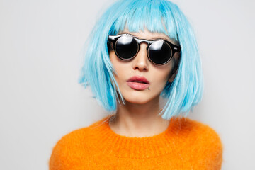 Studio close-up portrait of young girl with blue bob hairstyle, wearing sunglasses and orange sweater on white background.