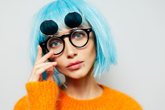 Close-up studio portrait of fashion girl with big blue eyes. Young woman wearing cyan wig, sunglasses and orange sweater on white background.