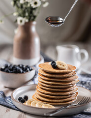 Stack of home baked banana pancakes with blueberries, gravy boat with maple syrup hangs over pancakes
