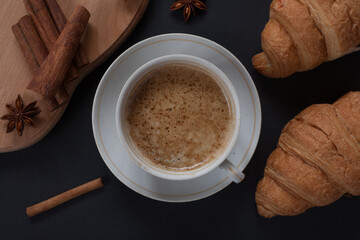 Cup of aromatic cappuccino with fresh croissant with cinnamon sticks on a dark background.