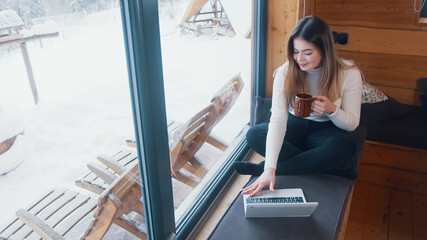 Young caucasian woman drinking coffee and having video call near the window. Turning laptop to show the snow. High quality photo