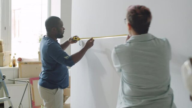 Afro-American man and his Caucasian wife measuring wall with tape and speaking while renovating home together