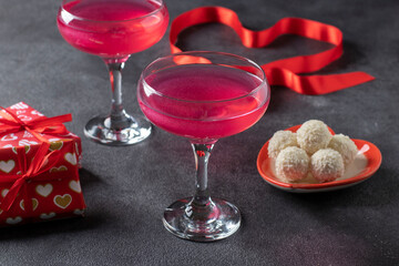 Two glasses of shimmering pink champagne, coconut candies in a heart-shaped plate and gifts against a dark background. Romantic dinner on Valentine's Day.