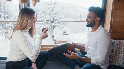 Happy multiracial couple drinking hot beverage and looking at snow outside. Winter holidays, Christmas or Valentines day. High quality photo