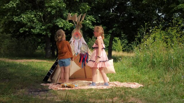 Two hippie sisters pillow fight outside. Boho girls having fun in nature near the wigwam. Children are parading the Indians.