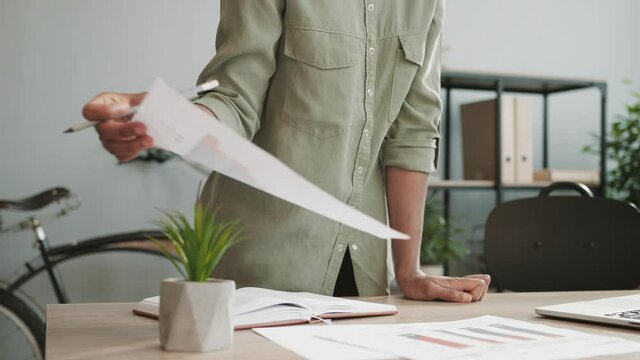 Young beautiful business lady with glasses standing over her desk looking through paper documents puts marks and signatures.