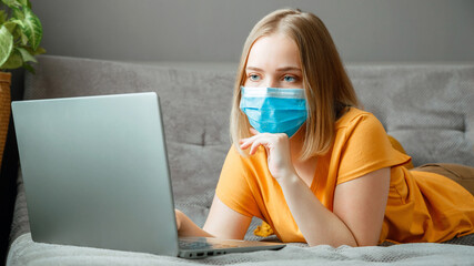 Woman in medical protective mask work using laptop at Home office lies on couch. Teen girl in mask Online learning education via laptop covid 19 lockdown time. Remote work Coronavirus pandemic banner