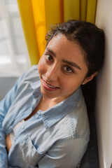 Portrait of a brunette woman sitting on a sofa in her room. Wear a denim shirt and black pants.