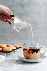 A woman's hand pours cream into black coffee. Coffee in a transparent cup, next to homemade cookies. Grey background.
