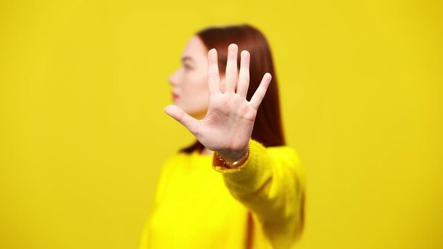 Close-up of female hand stretched to camera with blurred young redhead woman at yellow background turning away. Angry Caucasian lady making no gesture rejecting.