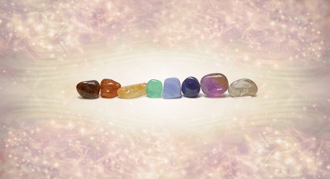 Single row of 8 chakra coloured healing crystals - Red, orange carnelian, yellow citrine, green, blue lace agate, indigo, ametrine & clear quartz, against pale peach sparkle background with copy space
