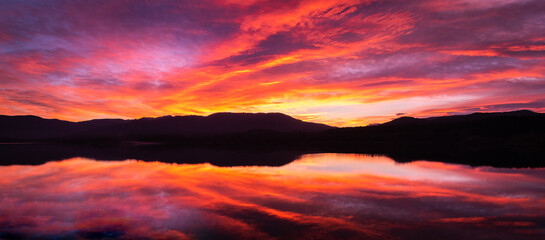 Sunrise or sunset with orange, purple and red clouds reflected in the lake water. Natural mirror at golden hour. Spectacular and beautiful scenery. Estany de Banyoles, Girona, Catalonia, Spain.